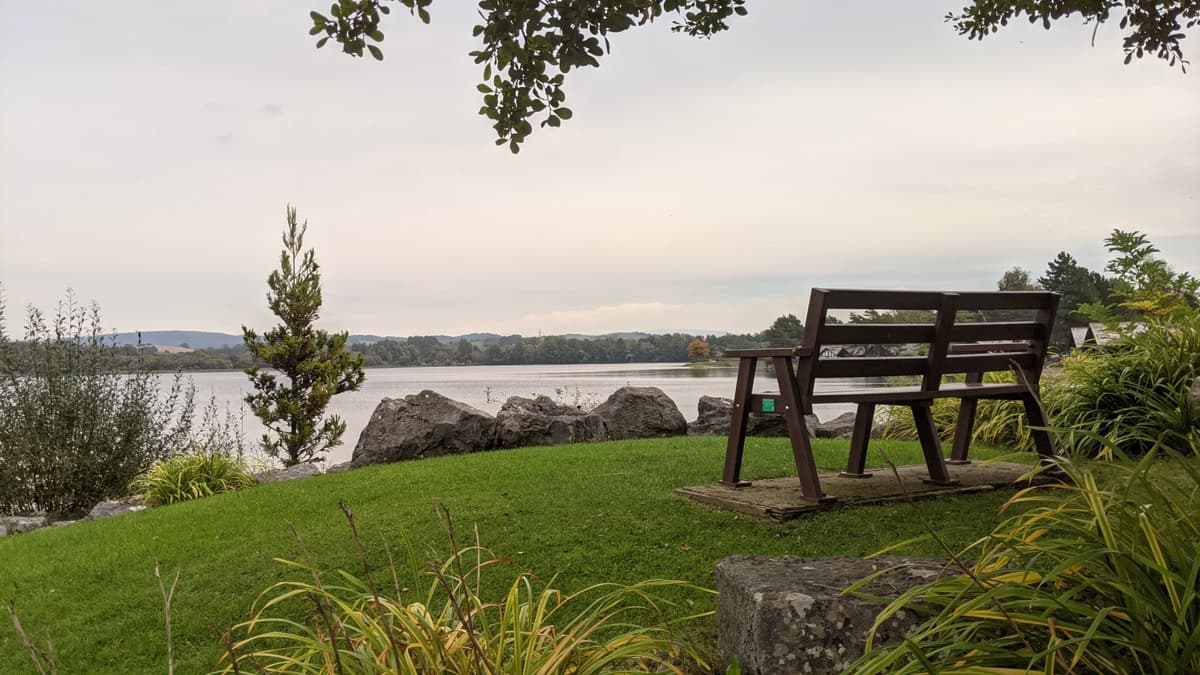A lakeside view with calm water, trees, and a wooden bench on well maintained bright green grass, facing the lake. The sky is overcast, and there is a hint of colour from the sun set, creating a quiet, peaceful atmosphere. Hills and forests can be seen in the far distance.