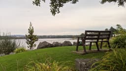 A lakeside view with calm water, trees, and a wooden bench on well maintained bright green grass, facing the lake. The sky is overcast, and there is a hint of colour from the sun set, creating a quiet, peaceful atmosphere. Hills and forests can be seen in the far distance.
