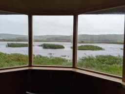 A view over a wide expanse of marshland seen through the windows of a wooden bird hide. The water is choppy and windswept, with reeds and small islands of vegetation under a grey, stormy sky.
