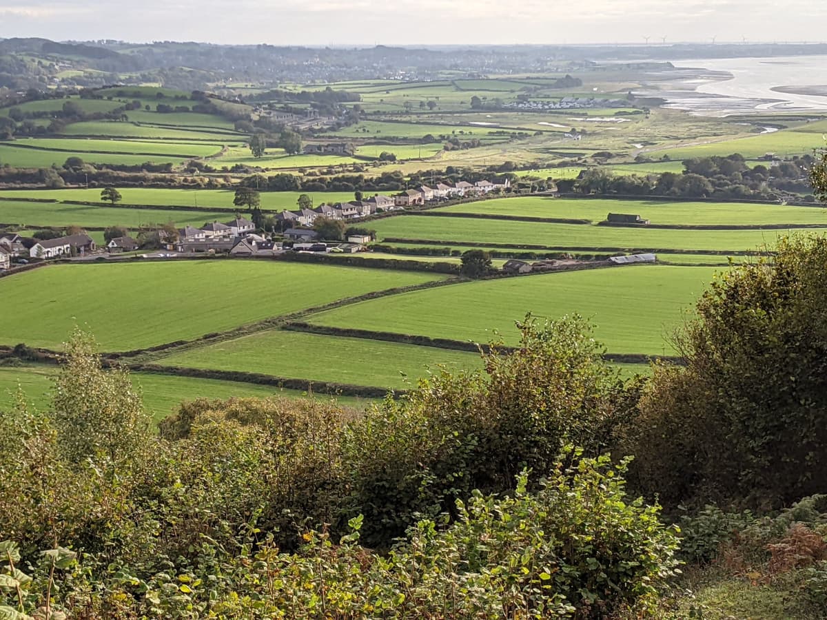A view from high up on a hill overlooking green fields, a small english village, rolling hills and the sea coming in at the top right. Everything looks miniature from this height. It is an overcast day but the light is shining through the clouds giving everything a golden hue.
