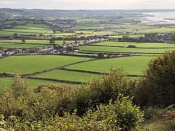 A view from high up on a hill overlooking green fields, a small english village, rolling hills and the sea coming in at the top right. Everything looks miniature from this height. It is an overcast day but the light is shining through the clouds giving everything a golden hue.