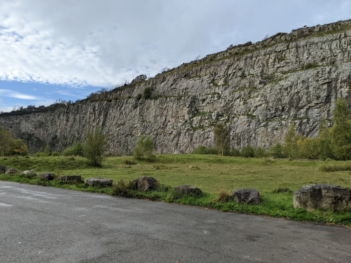A photo taken from the base of a tall limestone quarry. The sheer grey cliff rises dramatically with small patches of grass on the ledges. At the bottom, there’s a grassy area with a few trees, and the white cloudy sky shows bits of bright blue breaking through.